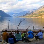 A group of six people sit together on a beach and look at the view of water and mountains.