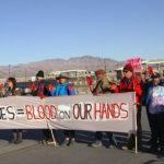 A group of people with bright red hands holding protest signs. The biggest reading, "Drones = Blood on our hands"